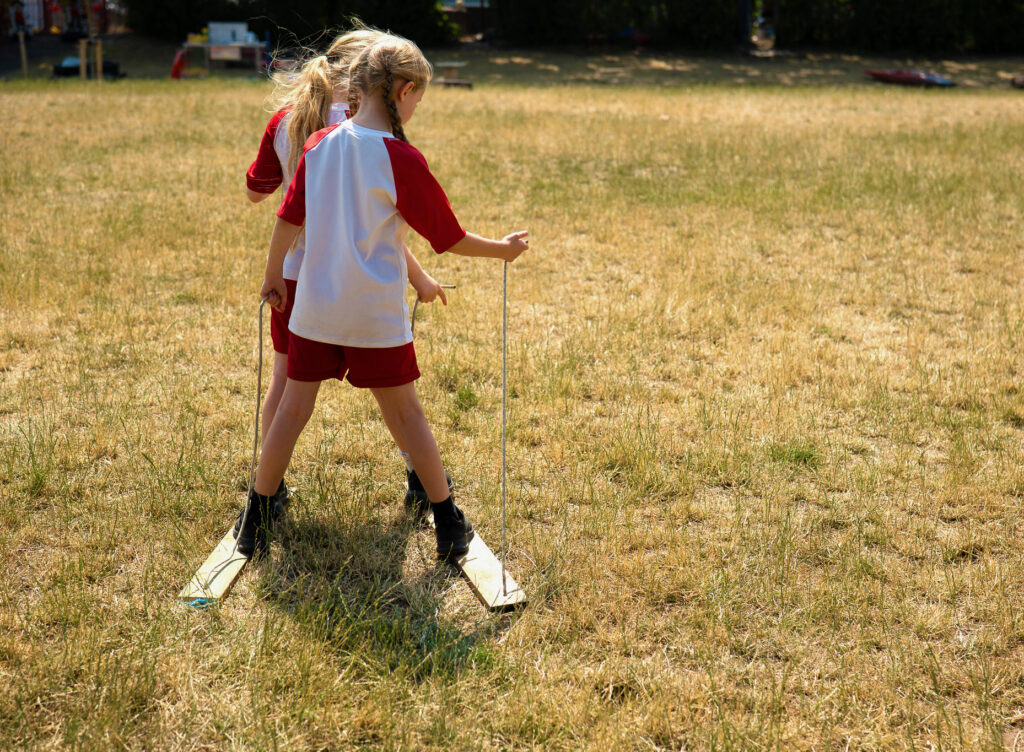 Two children in a field are standing with their feet on two wooden planks, holding onto rope that is connected to the plank.